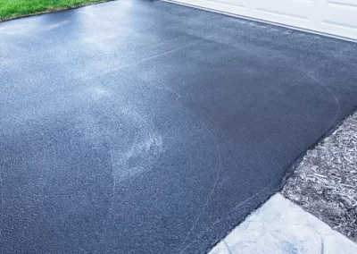 Freshly sealed black asphalt driveway by Asphalt Paving Experts in Middletown NY, beside a white garage door with a green lawn and trash bins nearby.