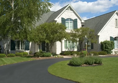 A large suburban house with white brick, green shutters, a tree, and a curved asphalt driveway paved by Asphalt Paving Experts shines under the blue sky in Middletown NY.