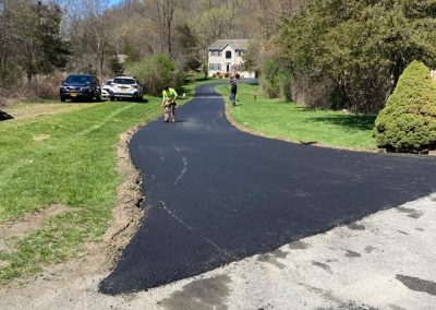 Workers from Action Paving are providing expert asphalt paving solutions, paving a driveway with fresh asphalt on a sunny day in Middletown NY, with cars and trees in the background.