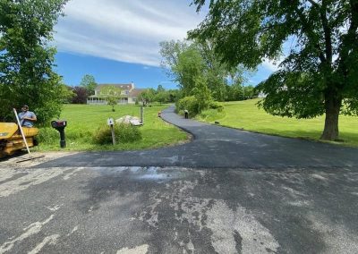 Freshly paved driveway by Asphalt Paving Experts leads to a house, surrounded by green grass and trees under a blue sky.