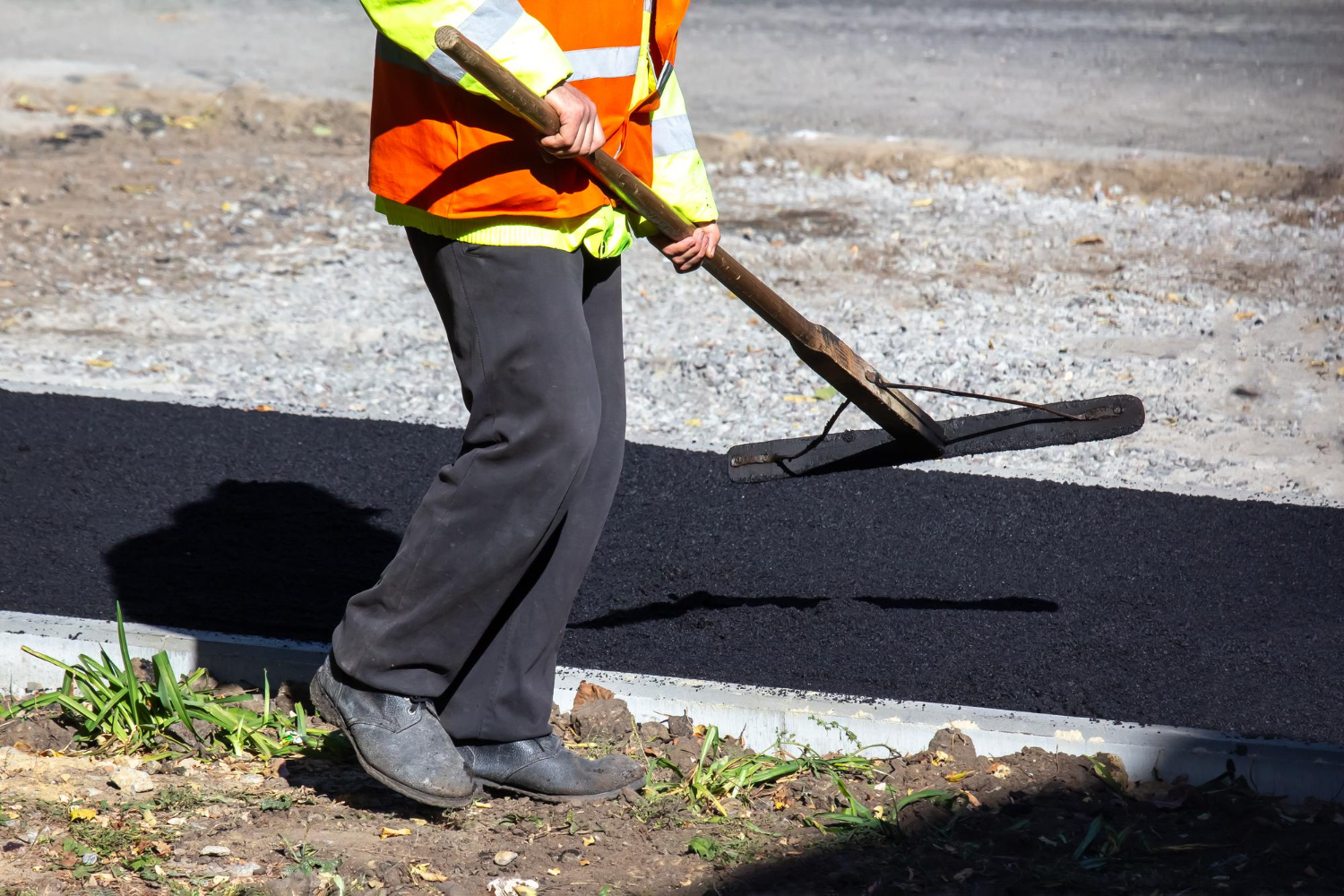 Worker in safety vest leveling fresh asphalt on a road with a wooden tool; only lower body visible.